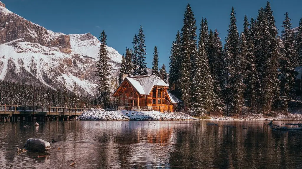 A wooden cabin, perfect for Airbnb management, is surrounded by snow-covered trees and reflected in a calm lake, with mountains and a clear blue sky in the background.