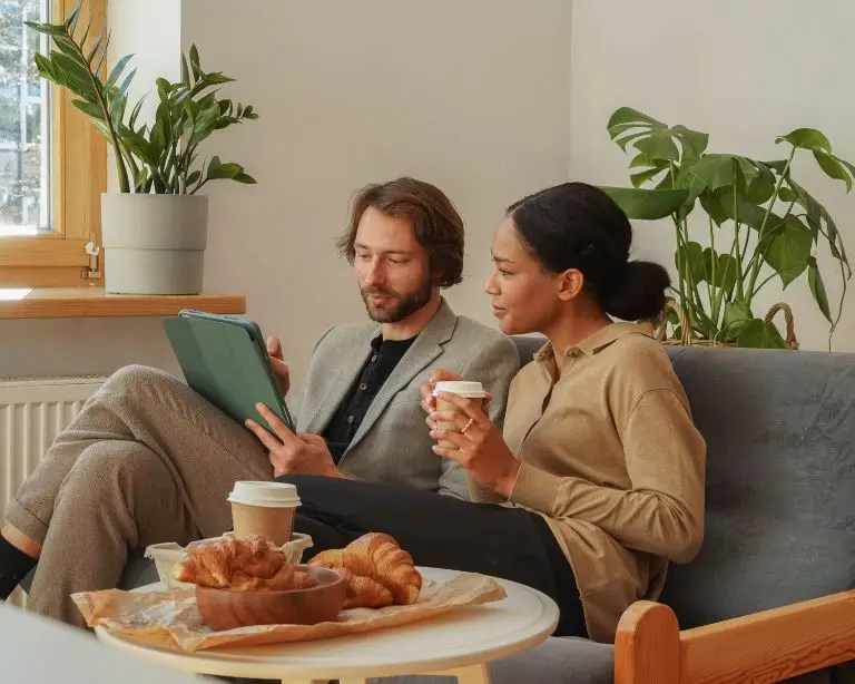 Two people sit on a couch with coffee and pastries, discussing the latest trends in short term rental management. One holds a tablet, likely browsing an Airbnb agency site. Lush indoor plants create a cozy atmosphere in the background.