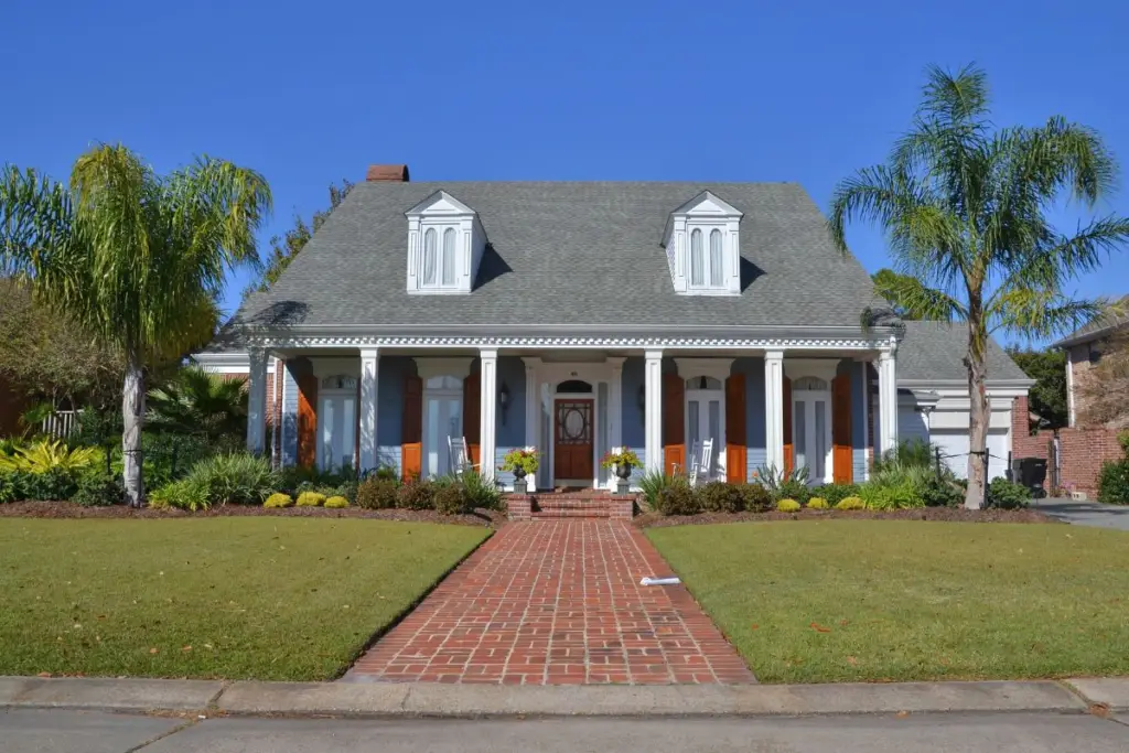 A charming two-story house with a gabled roof, white columns, and orange shutters is available through our Airbnb agency. It boasts a brick walkway, manicured lawn, and palm trees against a clear blue sky—a perfect escape booked effortlessly with our expertise in short-term rental management.