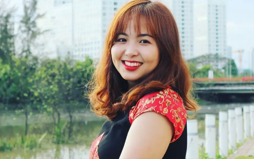 A woman with long brown hair smiles outdoors in a red patterned dress, perhaps enjoying a day off from her role at an Airbnb agency. Buildings and greenery frame the cheerful scene.