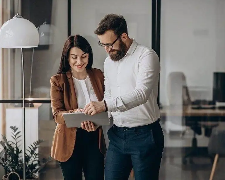 Two people, a man and a woman, stand in an office looking at a tablet together. The office has a modern design with glass walls and greenery.